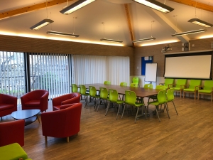 The Bradbury Meeting room for hire showing a well lit room with a boardroom table, projector screen and informal seating area.